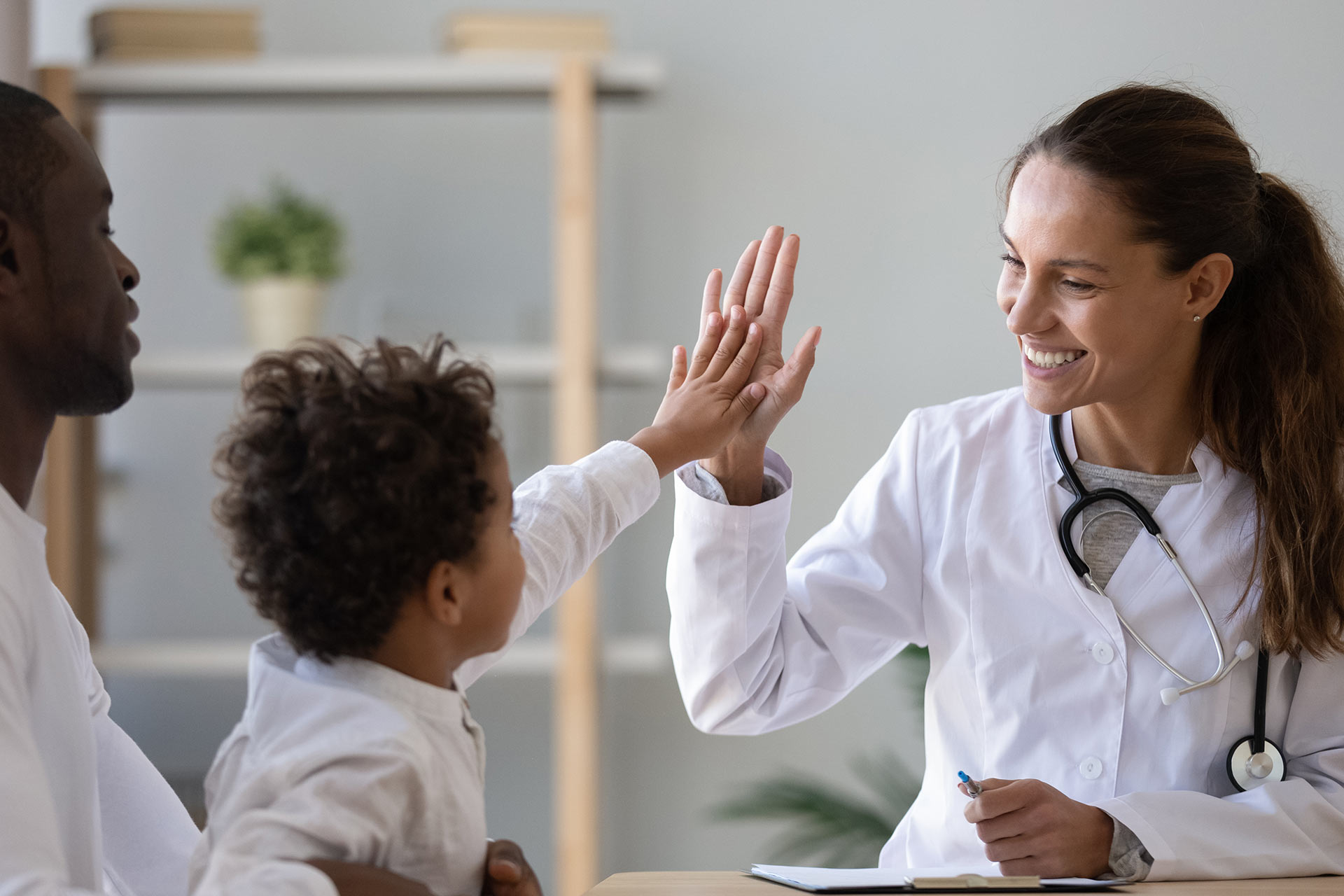 an image of a physician high fiving with her young patient while he sits on his dad's lap